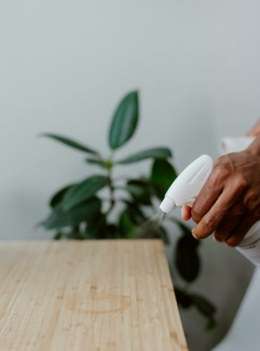 A hand using a spray bottle to clean a wooden table indoors, with a plant in the background.